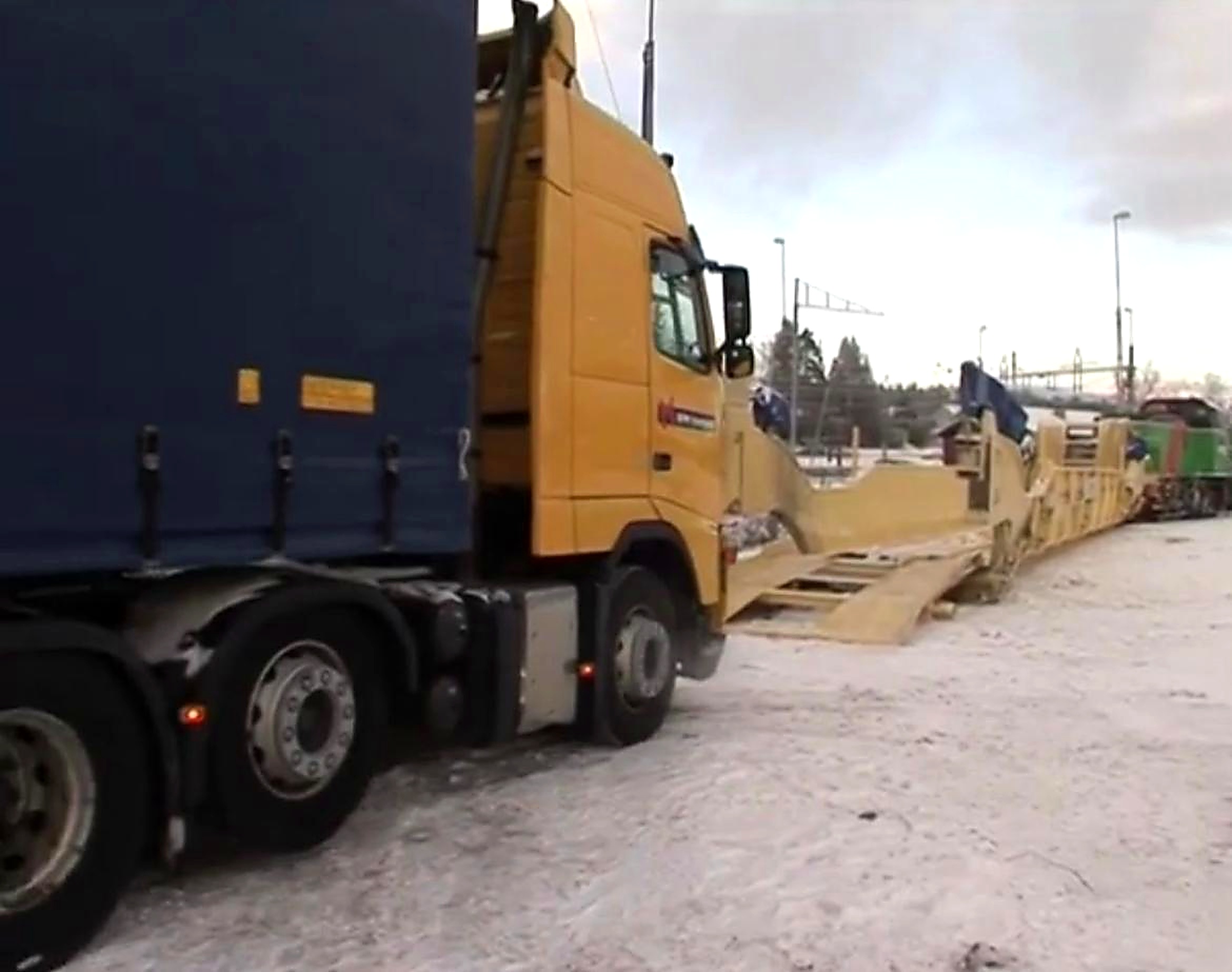 A yellow truck driving onto an Interwagon rail wagon via integrated ground-level ramps at a snowy rail siding